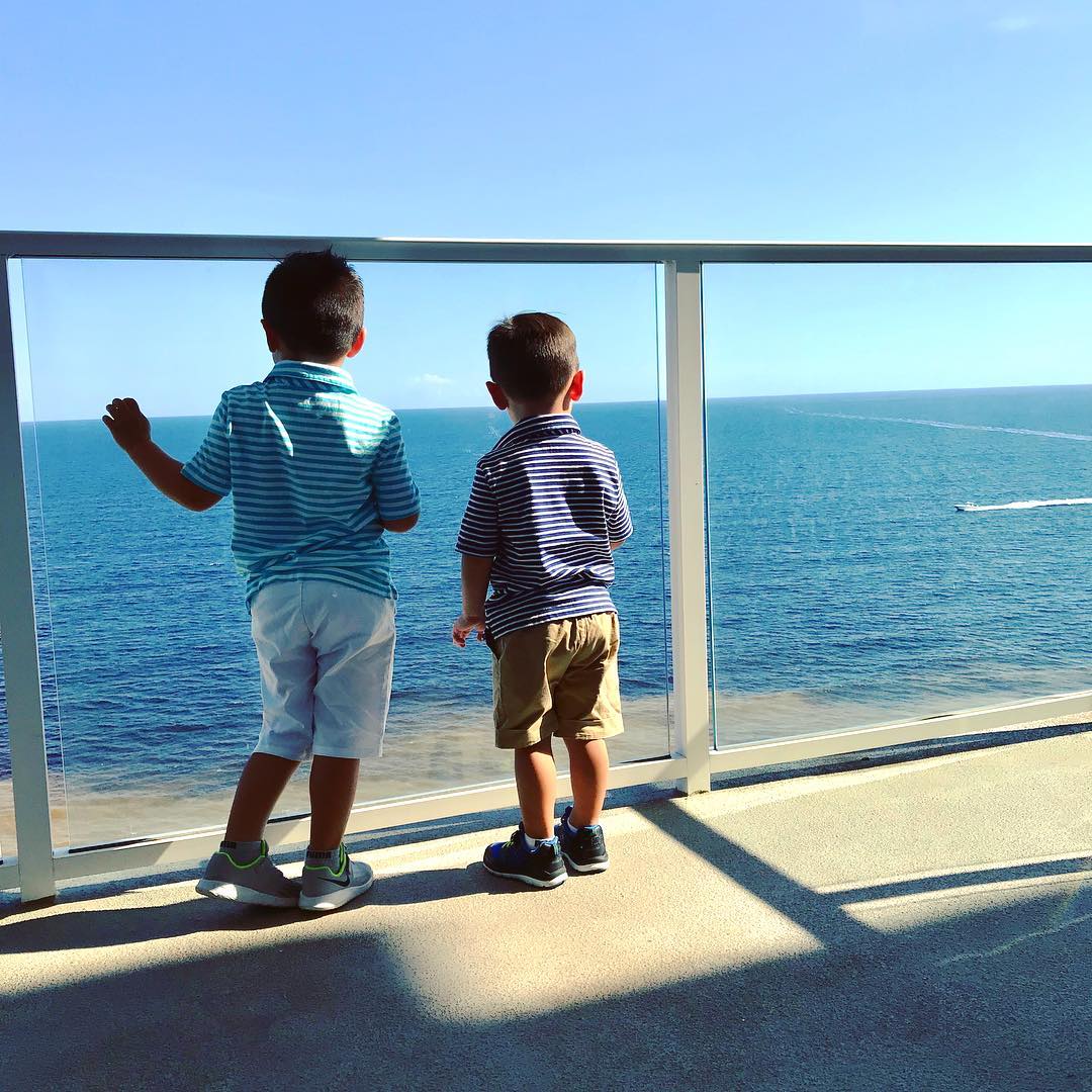 Two young boys stand on a balcony, peering through the railing to view the ocean.